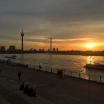 Die Sonne geht über einem Fluss unter, Menschen sitzen am Ufer und erleben ein Gefühl von Heimat.