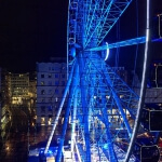 Ein großes, nachts beleuchtetes Riesenrad auf einem Stadtparkplatz.