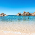 Ein Strand mit klarem Wasser und Sardinenfelsen im Hintergrund.