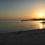 Die Sonne geht über dem Strand von Isola Rossa unter, auf dem sich Menschen befinden.