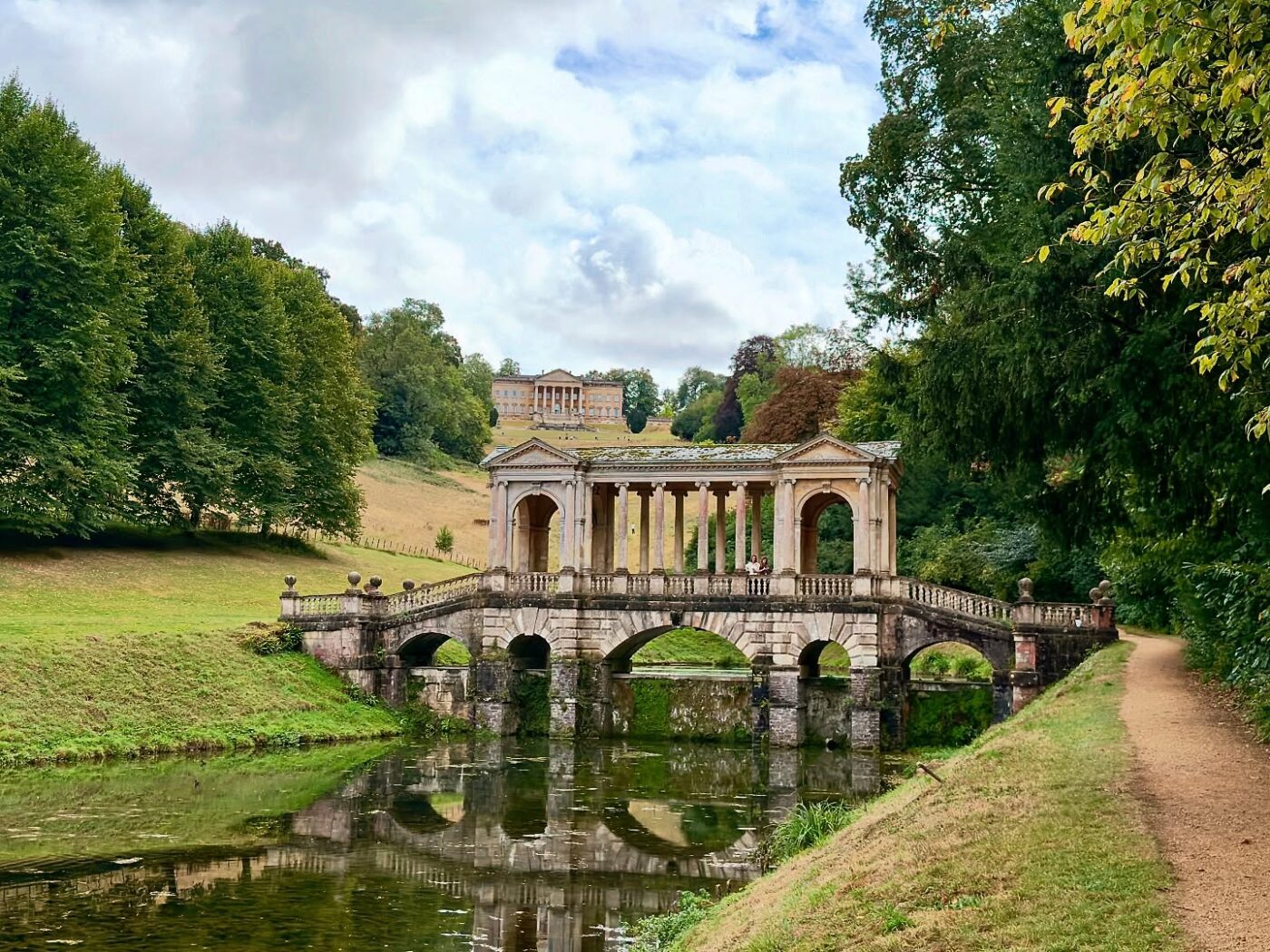 Eine historische Steinbrücke mit Säulen überspannt einen ruhig fließenden Fluss, umgeben von grünen Bäumen und Gras; im Hintergrund erhebt sich unter einem teilweise bewölkten Himmel ein großes, stattliches Gebäude auf einem Hügel.