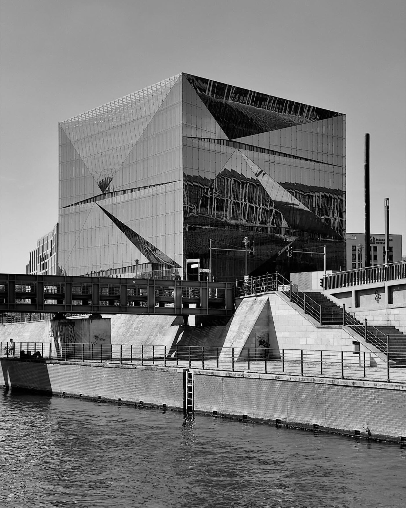 Am Wasser steht ein modernes Glasgebäude mit geometrischen Reflexionen, das sich durch kantige Architektur, Treppen und Geländer auszeichnet. Das Bild ist in Schwarz-Weiß gehalten.