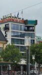 Modernes mehrstöckiges Gebäude mit der Aufschrift „AVALON“ in leuchtend orangefarbenen Buchstaben auf dem Dach, mehreren Flaggen, großen Fenstern und Balkonen, im Vordergrund Bäume und Straßenlaternen.