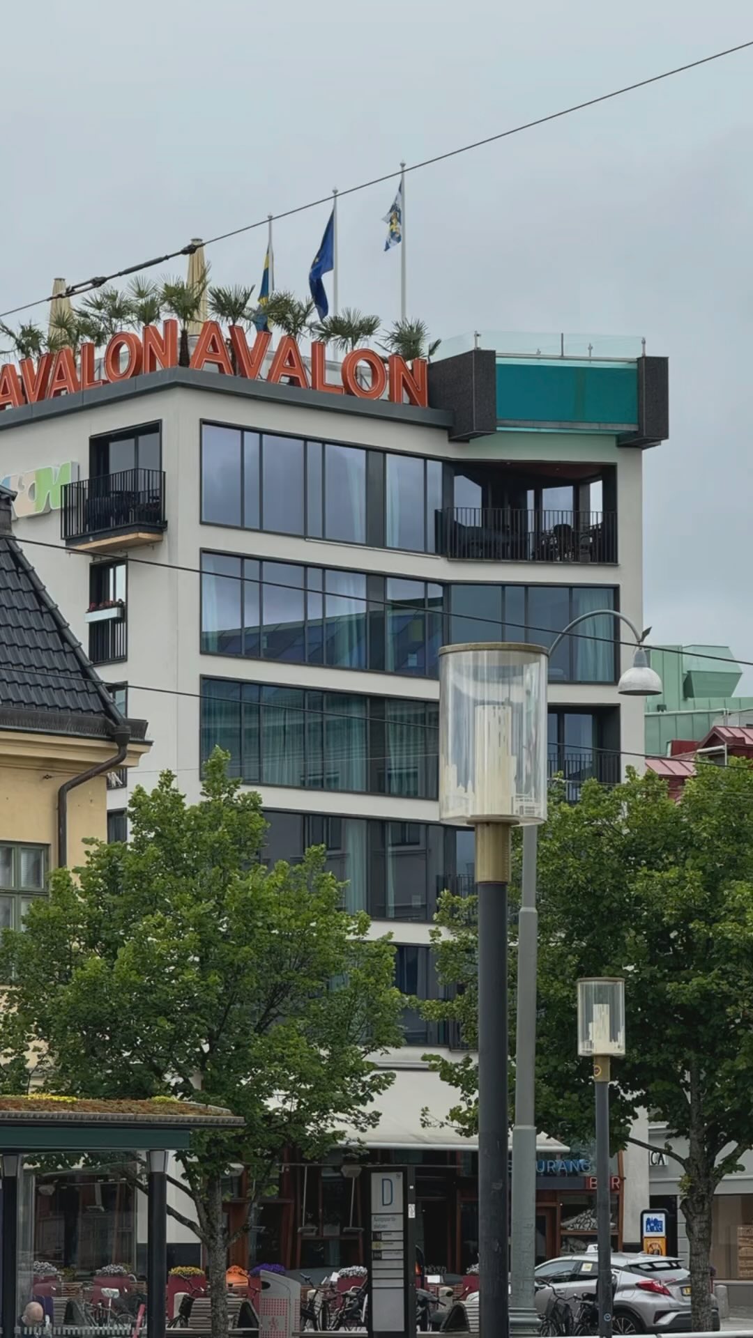 Modernes mehrstöckiges Gebäude mit der Aufschrift „AVALON“ in leuchtend orangefarbenen Buchstaben auf dem Dach, mehreren Flaggen, großen Fenstern und Balkonen, im Vordergrund Bäume und Straßenlaternen.