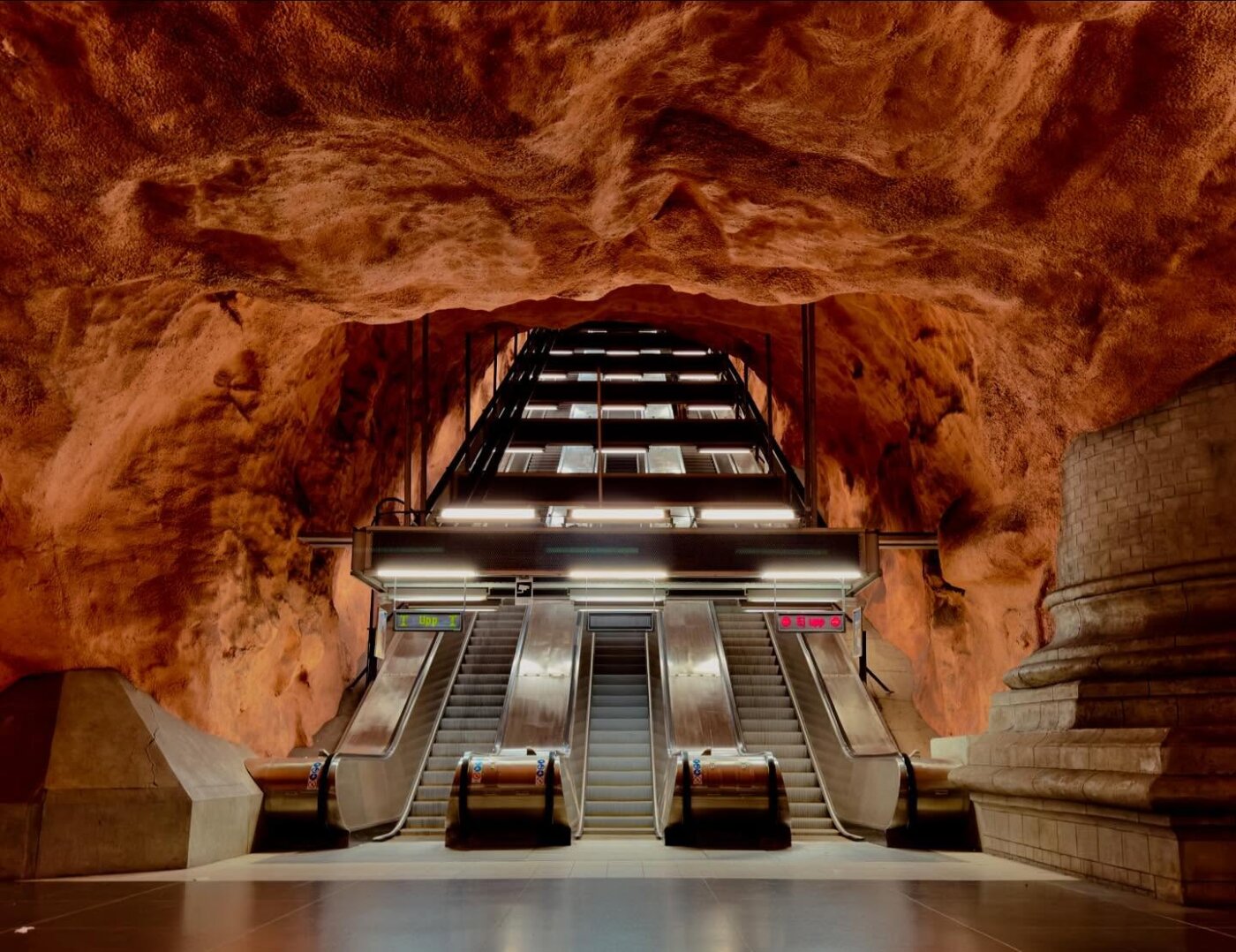 U-Bahn-Station mit Rolltreppen, umgeben von orangefarbenen, höhlenartigen Felswänden und einer entsprechenden Decke. Helle Deckenbeleuchtung erhellt den Bereich. In der Szene sind keine Menschen zu sehen.