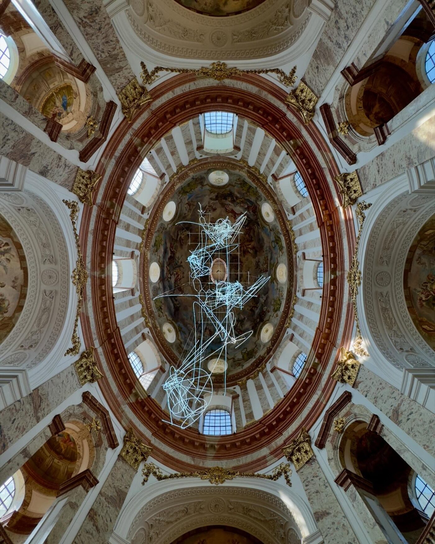 Eine kunstvoll gestaltete Kuppeldecke mit klassischen Fresken, architektonischen Details und einer modernen, abstrakten Lichtskulptur, die in der Mitte aufgehängt ist.