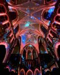 Bunte Lichter erhellen die Gewölbedecke und die Bögen im Inneren einer großen gotischen Kathedrale, wobei Blau- und Rosatöne eine dramatische, festliche Atmosphäre schaffen.