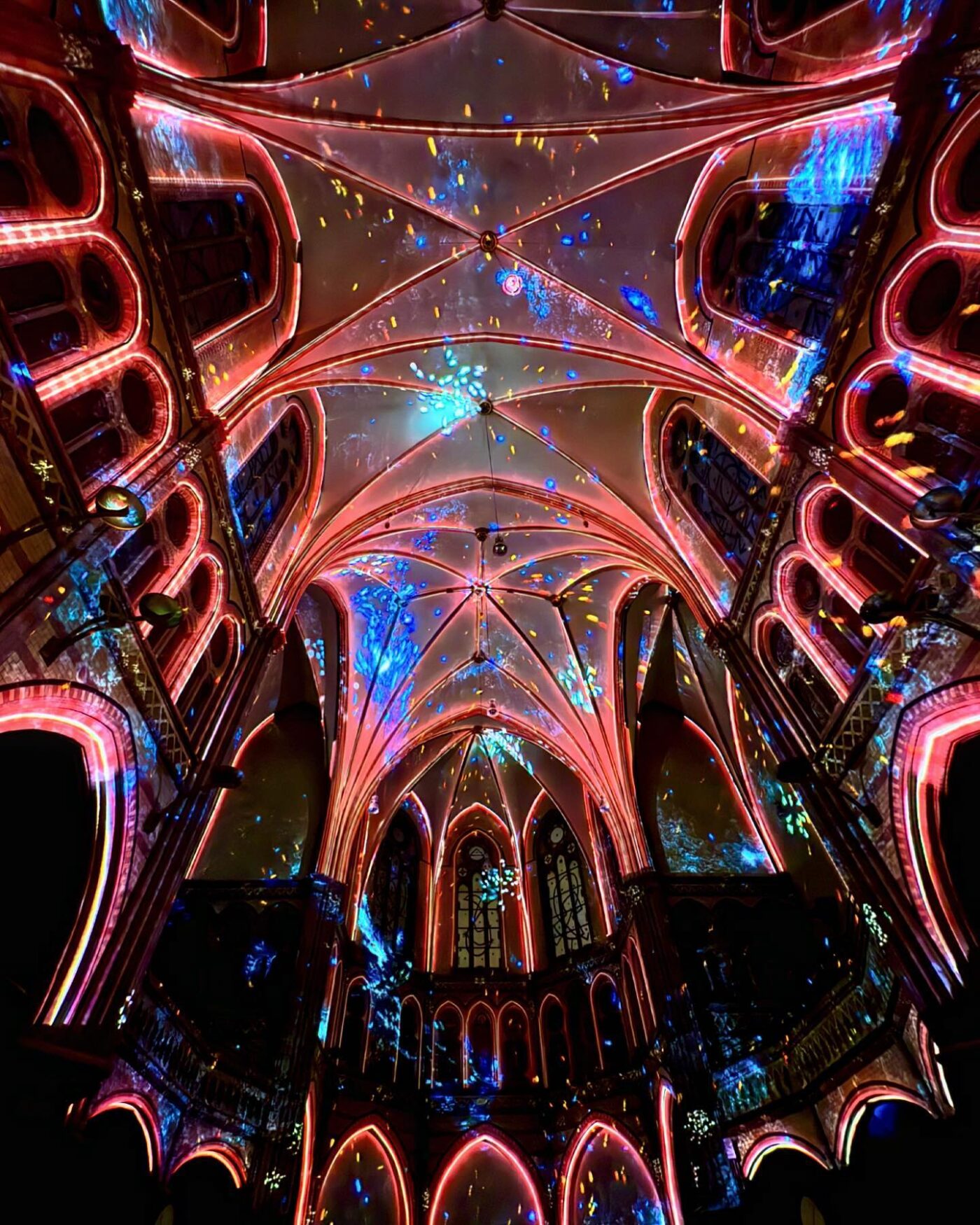 Bunte Lichter erhellen die Gewölbedecke und die Bögen im Inneren einer großen gotischen Kathedrale, wobei Blau- und Rosatöne eine dramatische, festliche Atmosphäre schaffen.