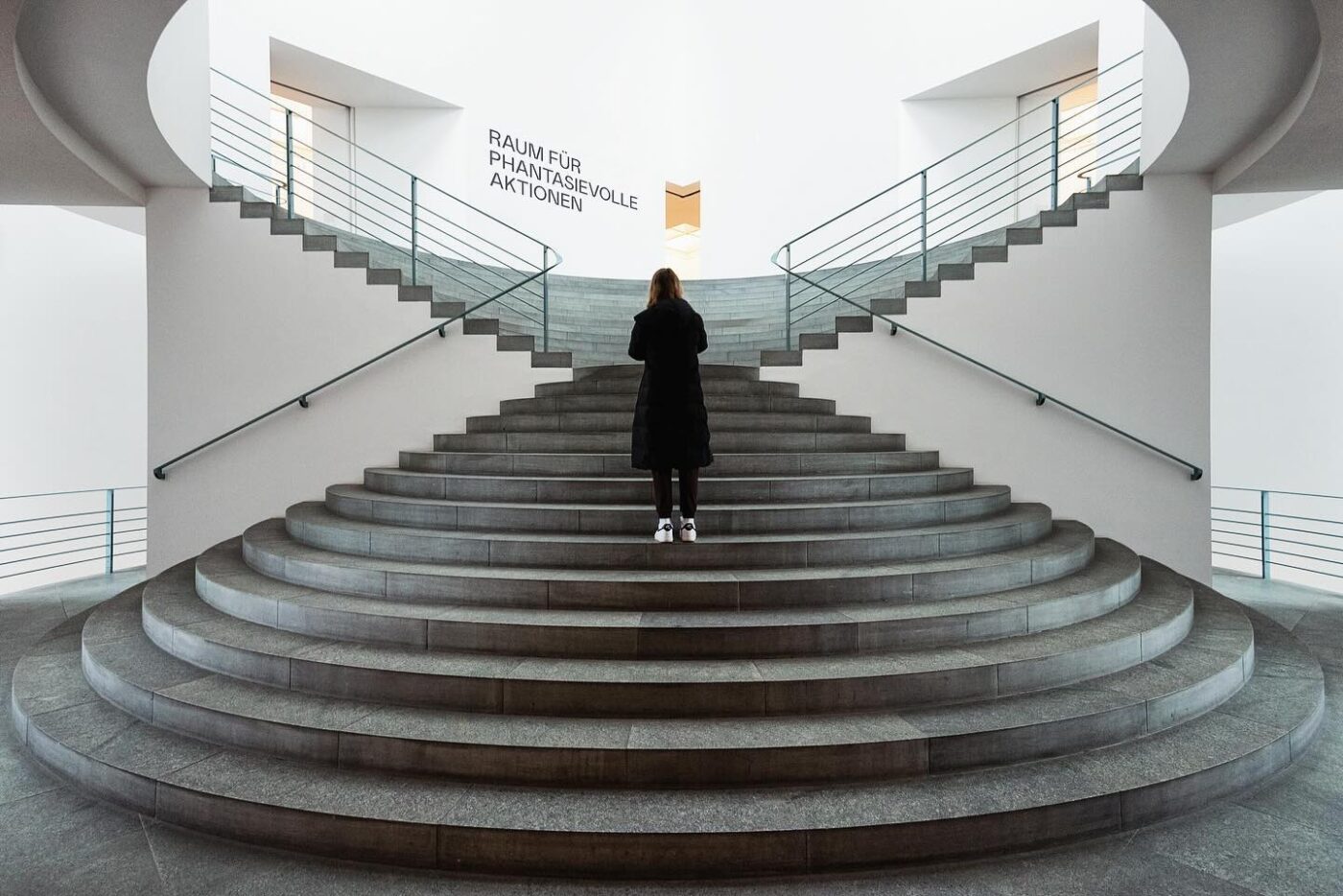 Eine Person in einem schwarzen Mantel steht am Fuße einer breiten, geschwungenen Treppe in einem modernen, minimalistischen Gebäude mit weißen Wänden und grauen Stufen.