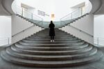 Eine Person in einem schwarzen Mantel steht am Fuße einer breiten, geschwungenen Treppe in einem modernen, minimalistischen Gebäude mit weißen Wänden und grauen Stufen.