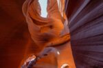 Ein Blick in einen schmalen Schlitzcanyon mit glatten, geschwungenen roten und orangefarbenen Felswänden, durch die das Sonnenlicht von oben hereinströmt.