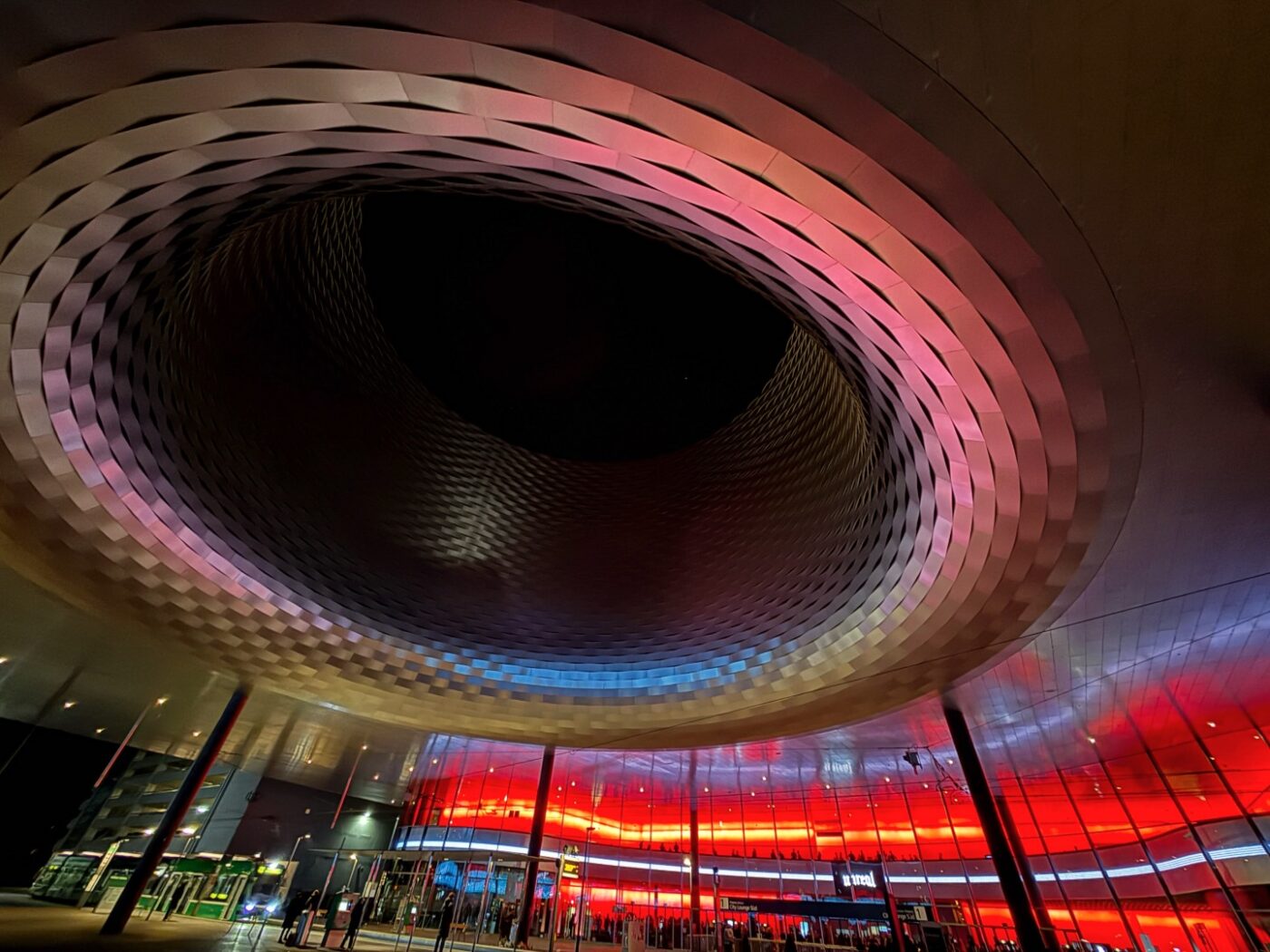 Rundes, modernes Gebäude mit einem großen offenen Innenraum, das nachts von roten und blauen Lichtern beleuchtet wird, mit Glastüren und Spiegelungen am Boden.