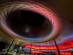 Rundes, modernes Gebäude mit einem großen offenen Innenraum, das nachts von roten und blauen Lichtern beleuchtet wird, mit Glastüren und Spiegelungen am Boden.