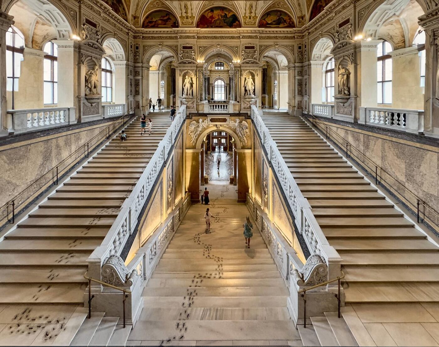 Eine prächtige Marmortreppe mit kunstvoll verzierten Geländern, Statuen und Bögen; Menschen, die hinauf- und hinuntergehen; große Fenster und Wandgemälde schmücken die Halle.
