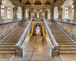 Eine prächtige Marmortreppe mit kunstvoll verzierten Geländern, Statuen und Bögen; Menschen, die hinauf- und hinuntergehen; große Fenster und Wandgemälde schmücken die Halle.