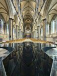 Innenansicht einer gotischen Kathedrale mit Gewölbedecken, Stuhlreihen, hohen Säulen, Buntglasfenstern und einem großen, spiegelnden Taufbecken in der Mitte.