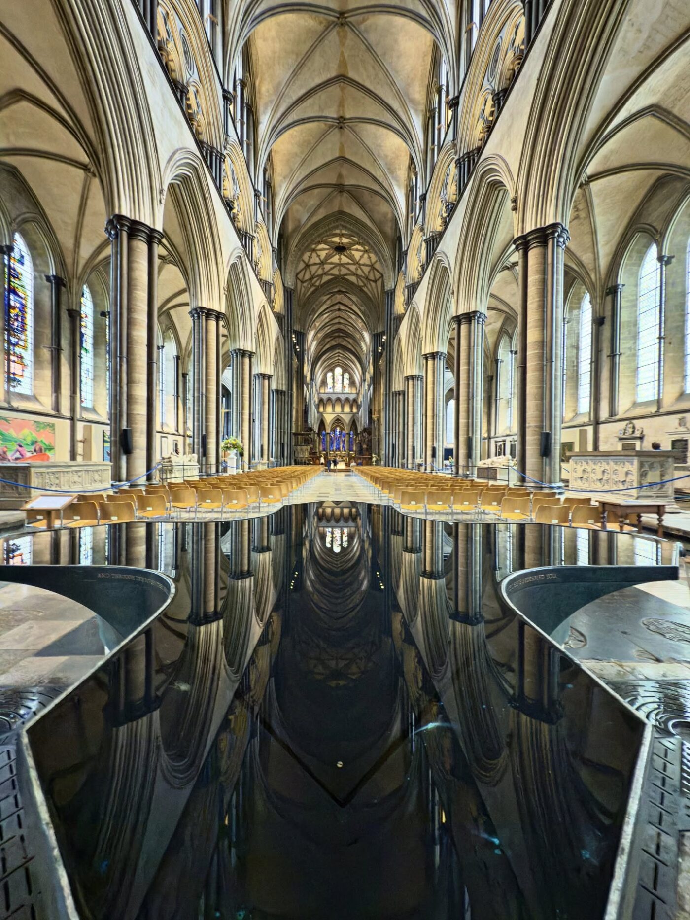 Innenansicht einer gotischen Kathedrale mit Gewölbedecken, Stuhlreihen, hohen Säulen, Buntglasfenstern und einem großen, spiegelnden Taufbecken in der Mitte.