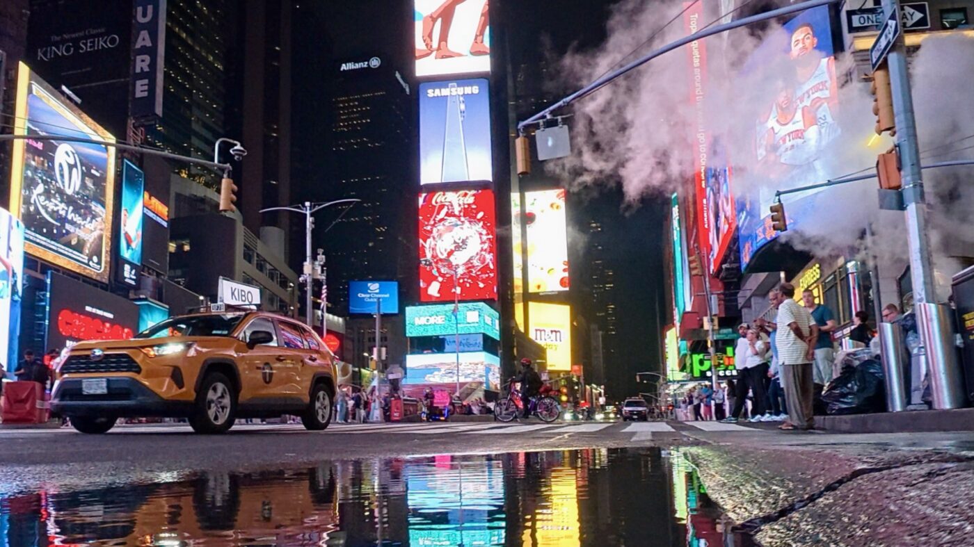 Ein gelbes Taxi fährt nachts durch den Times Square, vorbei an leuchtenden Werbetafeln, während Dampf aus einem Lüftungsschacht aufsteigt und sich die Lichter in einer Pfütze auf der Straße spiegeln.