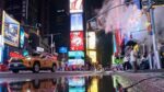 Ein gelbes Taxi fährt nachts durch den Times Square, vorbei an leuchtenden Werbetafeln, während Dampf aus einem Lüftungsschacht aufsteigt und sich die Lichter in einer Pfütze auf der Straße spiegeln.