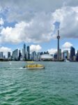 Ein gelbes Wassertaxi gleitet über das Wasser, im Hintergrund sind unter einem teilweise bewölkten Himmel die Skyline von Toronto und der CN Tower zu sehen.
