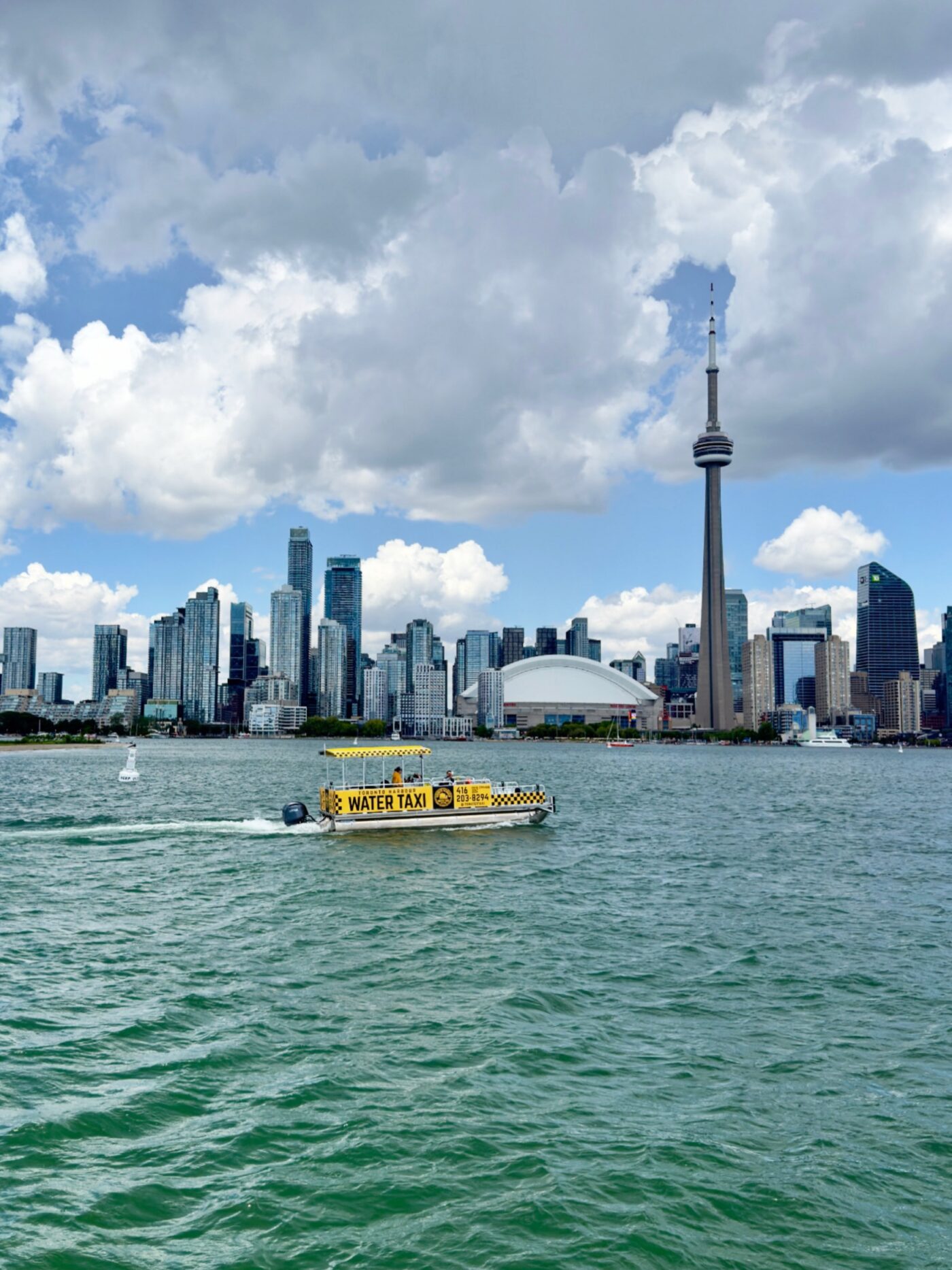 Ein gelbes Wassertaxi gleitet über das Wasser, im Hintergrund sind unter einem teilweise bewölkten Himmel die Skyline von Toronto und der CN Tower zu sehen.